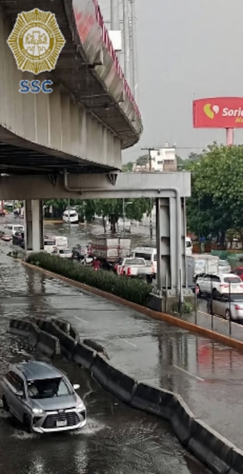 Carretera inundada con tráfico vehicular, bajo un viaducto elevado. Un vehículo atraviesa el agua. Barreras de concreto, señal de Soriana y logo SSC visible