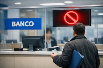 Hombre de espaldas con documento y carpeta azul ante ventanilla de banco con pantalla que muestra símbolo de prohibido y logo Banco