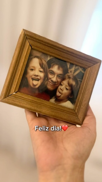 Una mano sostiene un marco de madera con una fotografía en blanco y negro de un hombre con gafas y dos niños. Todos sonríen y sacan la lengua. Aparece el texto "Feliz día!"