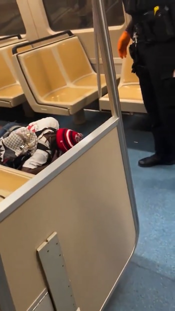 Un hombre con gorro rojo y blanco yace en el suelo de un vagón de tren MARTA con asientos beige. Un oficial de uniforme oscuro y guantes naranjas se acerca
