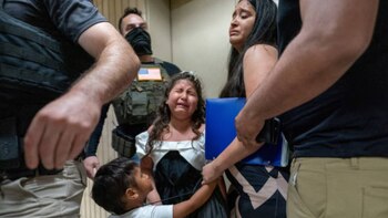 Niña con vestido blanco y tiara llora junto a mujer y niño. Hombres con chalecos tácticos y uno con bandera de EE. UU. los rodean, en un interior con paredes claras