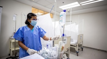 Mujer con mascarilla, gafas y bata azul ajusta el tubo de una bolsa de suero intravenoso junto a camas de hospital.