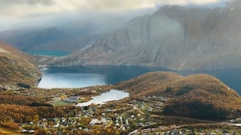 Vista aérea de un valle noruego con un pueblo, dos lagos, montañas escarpadas y bosques con follaje otoñal. El cielo está parcialmente cubierto de nubes