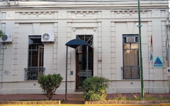 Fachada de un edificio blanco con ventana, puerta con toldo, dos pequeños arbustos y un cartel del Ministerio Público de la Acusación