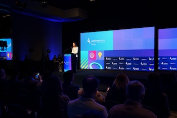 Una mujer habla desde un atril en un escenario. Detrás, pantallas muestran logos de AmCham Energy Forum y AmCham Argentina. Una audiencia se sienta de espaldas