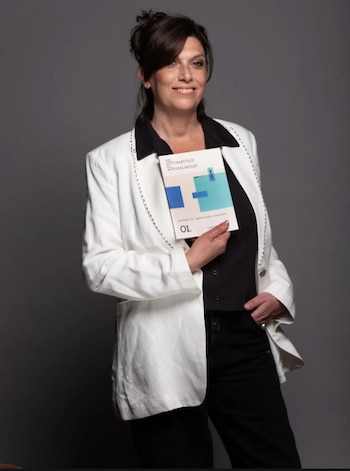 Mujer de cabello oscuro y blazer blanco sonríe, sosteniendo un libro blanco con título "Del Autosabotaje al Autosalvataje" en su mano izquierda, fondo gris