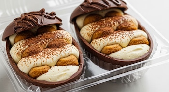 Vista de cerca de dos mitades de huevos de chocolate abiertos, rellenos de crema blanca, bizcochos y cacao en polvo, dentro de un recipiente transparente de plástico.