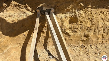 Excavación profunda en tierra arenosa con tres tablones de madera apoyados diagonalmente contra sus paredes. El suelo es de color marrón claro y hay sombras