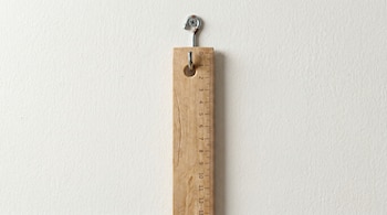 Regla de madera vertical con marcas de medición del 0 al 13, colgada de un gancho metálico sujeto a una pared blanca texturizada.
