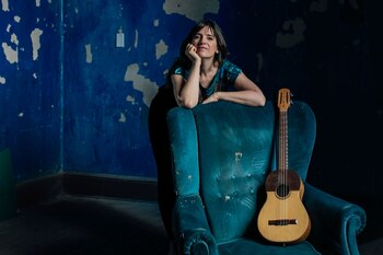 Mujer con camiseta estampada, apoyada en el respaldo de un sillón de terciopelo azul. Un instrumento de cuerda de madera clara se apoya en el sillón. Fondo azul