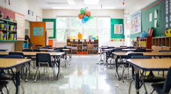 Interior de un aula de escuela vacía con pupitres y sillas alineados. Ventanas grandes, una pizarra verde y estantes llenos de materiales