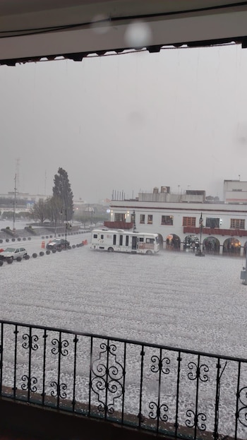 Vista desde un balcón de una plaza pública y calles cubiertas completamente de granizo. Se aprecian coches, un autobús blanco y edificios bajo un cielo nublado