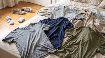 Cuatro camisetas arrugadas de colores neutros reposan sobre una cama deshecha, con un par de zapatillas y calcetines en el suelo.