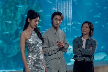 Tres actores posan frente a un fondo azul que simula un acuario con peces y patrones de luz; la mujer de la izquierda lleva un vestido plateado brillante