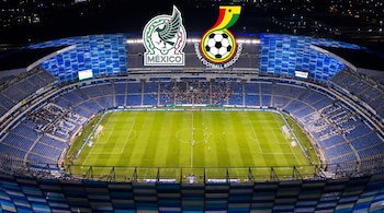 Vista aérea nocturna de un estadio de fútbol azul iluminado con un partido en curso. Logotipos de México y Ghana flotan sobre el campo.