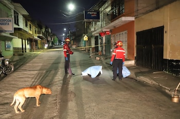 Dos miembros de servicios de emergencia, un perro de color marrón, un objeto cubierto con tela azul, cinta amarilla en una calle de noche con edificios