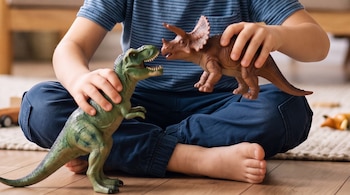 Un niño sentado en el suelo sostiene dos figuras de dinosaurios: un Tiranosaurio Rex verde y un Triceratops marrón, enfrentados.