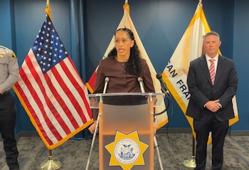 Brooke Jenkins habla tras un podio con el sello de la fiscalía de San Francisco, flanqueada por la bandera de Estados Unidos y dos personas
