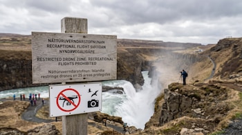 Un cartel de madera prohíbe el uso de drones recreativos y muestra símbolos de aves y cámara, ubicado frente a una gran cascada en Islandia con personas.