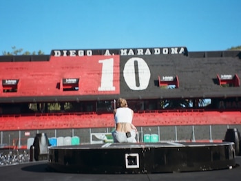 Vista trasera de Tini Stoessel sentada en un escenario circular con el estadio Diego Maradona de fondo, mostrando tribunas rojas y negras con el número 10
