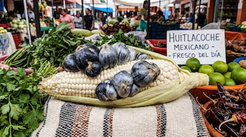 Un elote blanco con huitlacoche gris y negro, chiles secos rojos, cilantro, limas verdes y un letrero de 'Huitlacoche Mexicano' en un puesto de mercado.