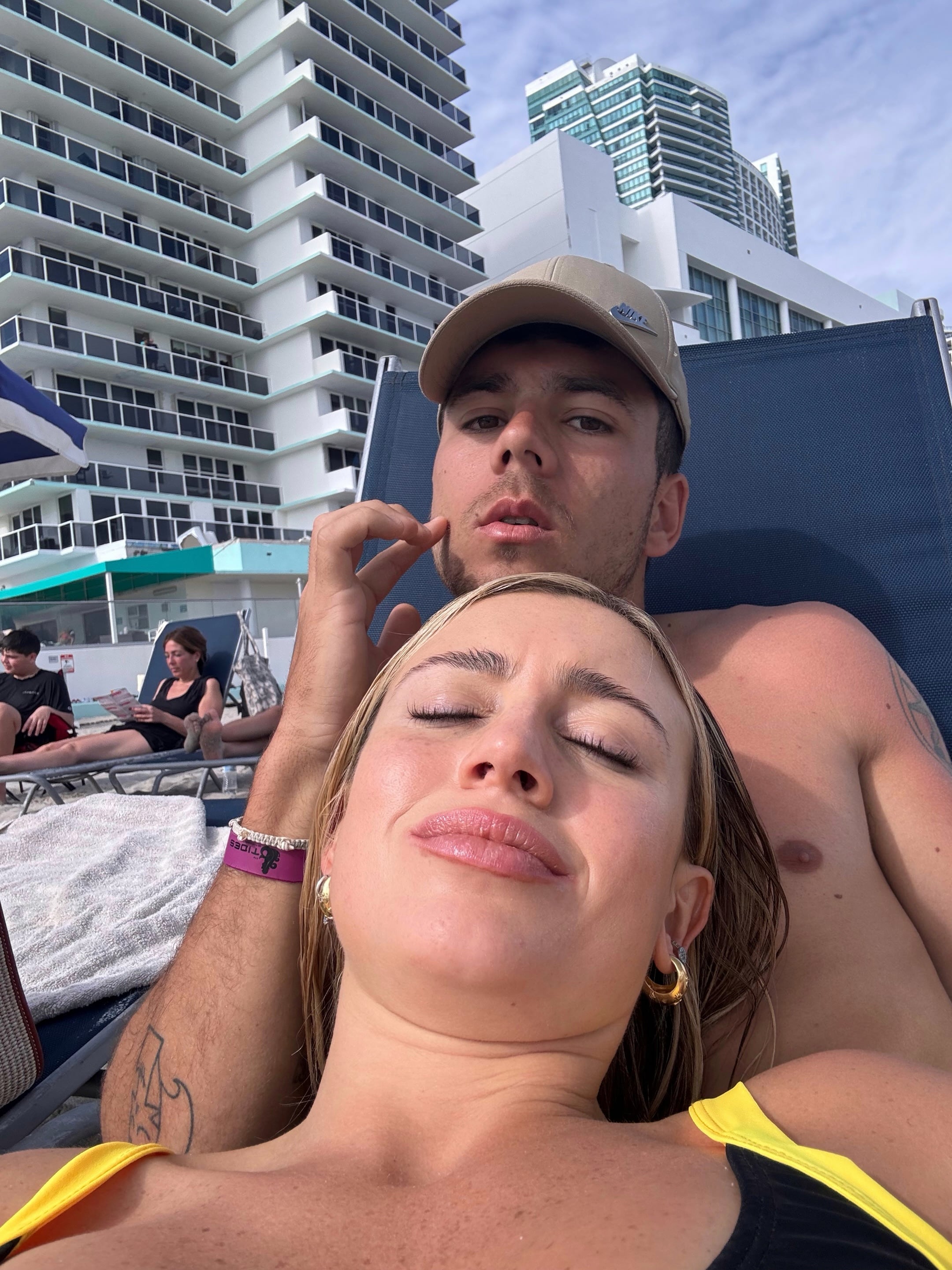 La periodista Morena Beltrán y el futbolista Lucas Blondel descansan en reposeras frente al mar con edificios de fondo.