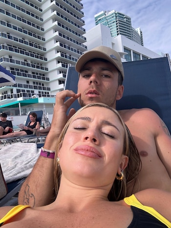 Un hombre con gorra beige y una mujer de cabello rubio con los ojos cerrados reposan en una silla de playa junto a grandes edificios