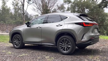 Un SUV compacto de color gris plateado con rines oscuros está estacionado en un camino de grava, con árboles y un muro de piedra de fondo