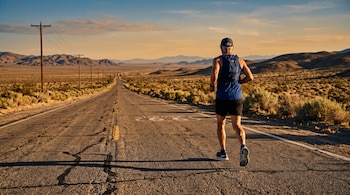 Un hombre de espaldas corre por una carretera asfaltada con la marca de la Ruta 66 rodeada de colinas desérticas.