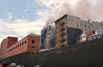 Un edificio de ladrillo oscuro emite humo denso. Otros edificios, una pared, vehículos y personas en la calle bajo un cielo nublado