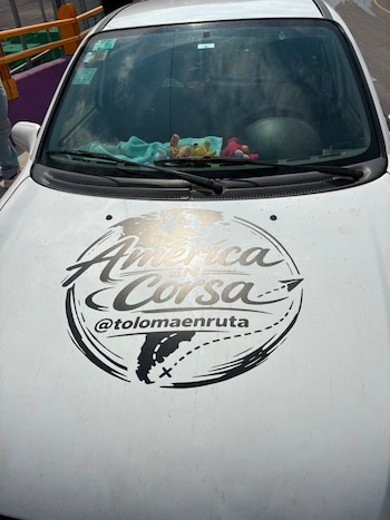 Un auto Chevrolet blanco con calcomanía "América en Corsa" y matrícula argentina está estacionado frente a un estadio con banderas del Mundial 2026