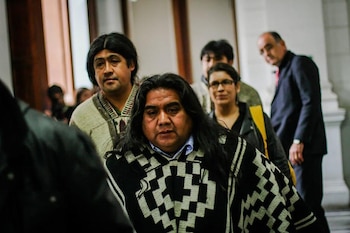 Un hombre con cabello largo y un poncho Mapuche de patrón geométrico blanco y negro mira al frente, rodeado por otras personas borrosas en un interior