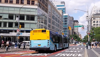 Un autobús azul y amarillo circula por una calle urbana rodeada de edificios, semáforos y personas en las aceras
