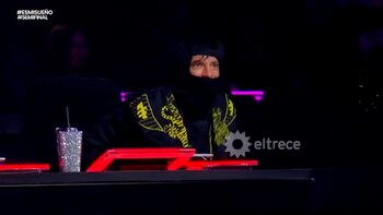 Un hombre con pasamontañas negro y chaqueta negra con diseños amarillos está sentado frente a una consola con luces rojas, con un vaso brillante y pajita