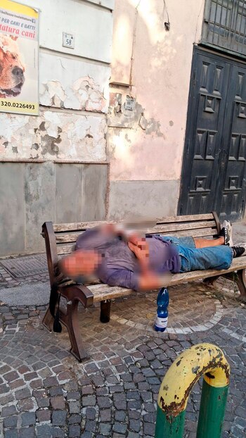 Un hombre yace recostado en un banco de madera. A su lado hay una botella de agua azul. Detrás, una pared con pintura descascarada y una puerta metálica