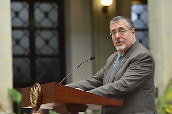 Hombre de mediana edad con traje gris, corbata azul y gafas, hablando en un atril de madera con micrófono, fondo interior borroso