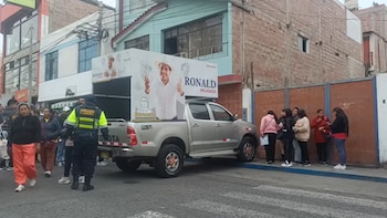 Una camioneta plateada con un panel publicitario de Ronald Hilasaca. Varias personas, un agente de seguridad municipal, edificios y una calle