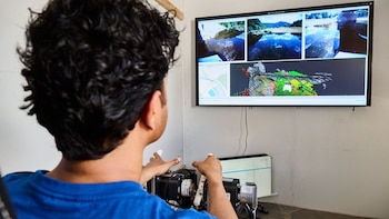 Hombre de espaldas, cabello rizado, camiseta azul. Observa pantalla grande con cuatro paneles: imágenes aéreas de un río, un mapa y datos 3D de terreno. Manos en controles