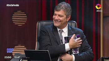 Mario Pergolini viste traje y corbata mientras abraza una caja rectangular en un estudio de televisión