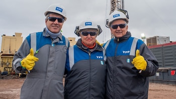Tres personas con cascos de YPF y ropa de trabajo. Un hombre, una mujer y otro hombre. Al fondo, un equipo de perforación y estructuras industriales bajo cielo nublado