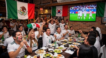 Grupo de aficionados mexicanos en un restaurante, vistiendo camisetas blancas y con caras pintadas, celebrando mientras ven un partido de fútbol en TV.