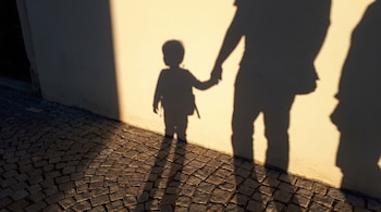 Las sombras de un niño y un adulto tomados de la mano se proyectan sobre una pared de color claro y un suelo de adoquines.
