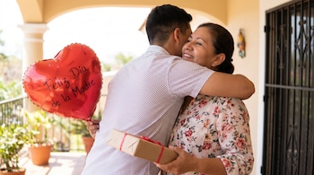 Un joven con camisa estampada abraza a una mujer sonriente con blusa floral, quien sostiene un regalo y un globo rojo en forma de corazón.