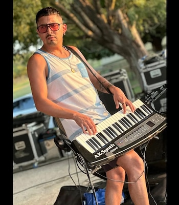 Un hombre con anteojos de sol y camiseta de tirantes toca un teclado colgado al hombro cerca de cajas de equipos