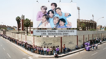 Un panel con la imagen de BTS sobre una acera donde varias personas hacen fila junto al muro exterior de un estadio.