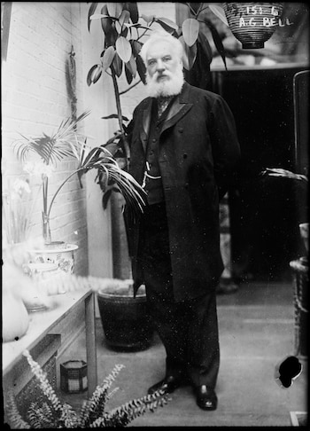 Fotografía en blanco y negro de Alexander Graham Bell, un hombre mayor con barba y cabello blancos, de pie en un traje oscuro entre varias plantas en macetas