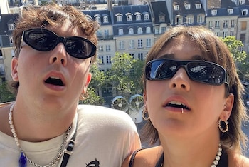 Primer plano de dos personas con gafas de sol, un hombre con collar de perlas y cadena metálica, una mujer con piercing labial, con edificios y árboles detrás