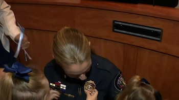 Una oficial de policía con uniforme oscuro se inclina mientras dos niñas, vistas de espaldas, tocan o miran su insignia. Fondo de pared de madera