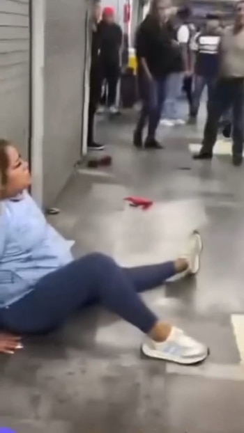 Una mujer con camiseta azul y leggings oscuros se encuentra sentada sobre el suelo de un pasillo. Varias personas y un objeto rojo son visibles en el fondo
