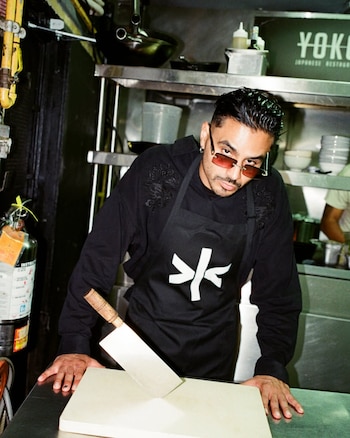 Álvaro Díaz, hombre con barba y gafas de sol rojas, en una cocina industrial. Viste sudadera negra y delantal, con un cuchillo grande sobre una tabla de cortar